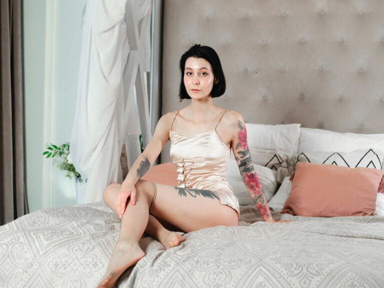 Tattooed woman in satin nightwear posing on an elegant bed with pillows and headboard.